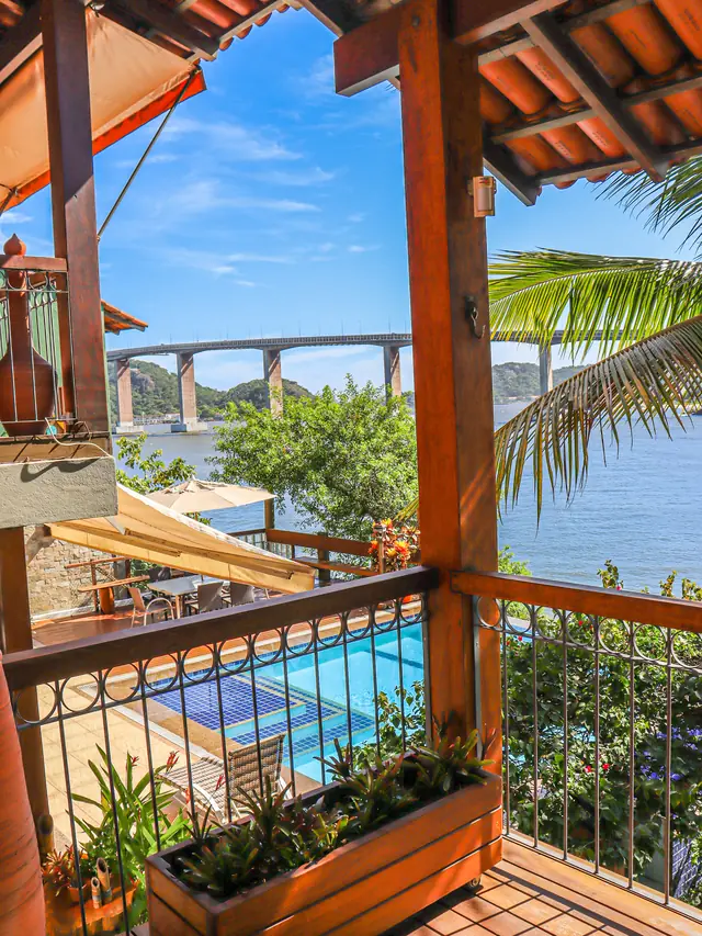 Vista da varanda da casa: colunas de madeira, a piscina abaixo, a baía de Vitória e a Terceira Ponte ao fundo