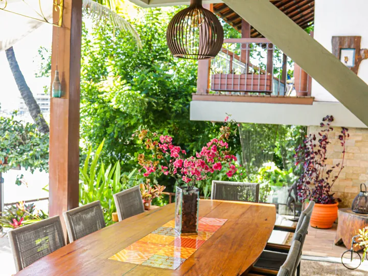 Mesa comprida de madeira no espaço gourmet coberto, com cadeiras dos dois lados e plantas em volta